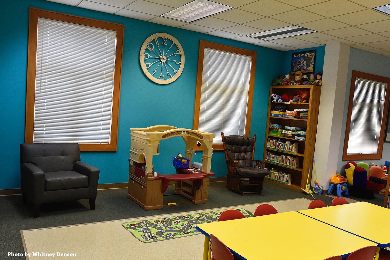 Children's play area with teal walls, windows, playhouse, shelves of toys, and yellow tables.
