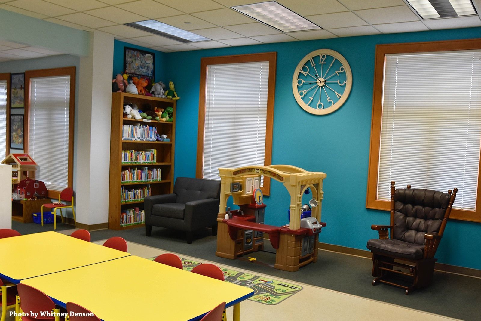 Library corner: yellow tables, bookshelves, rocking chair, toy playhouse against teal wall, and two windows with blinds.