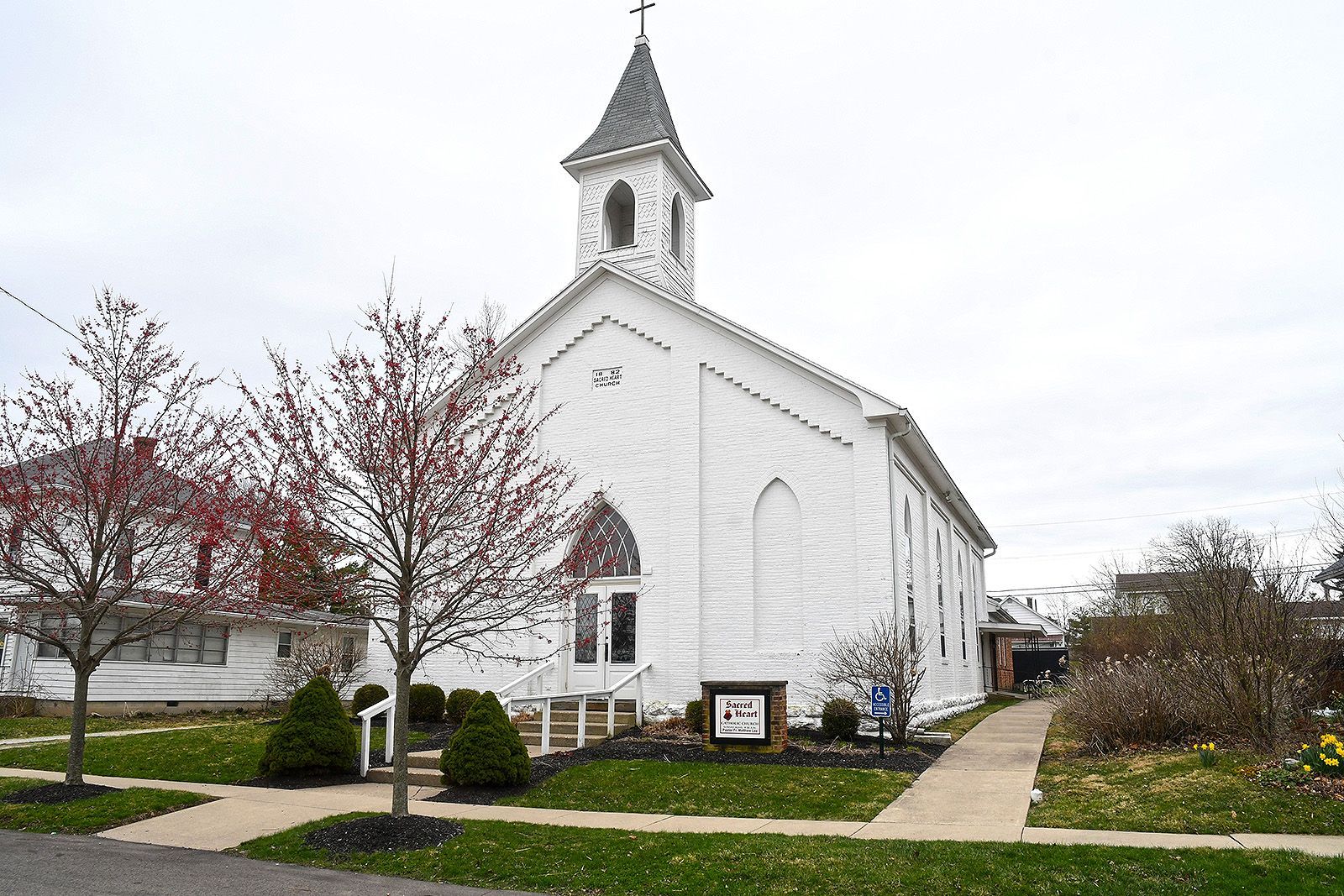 White church building with steeple, cross, and sign, on a cloudy day.