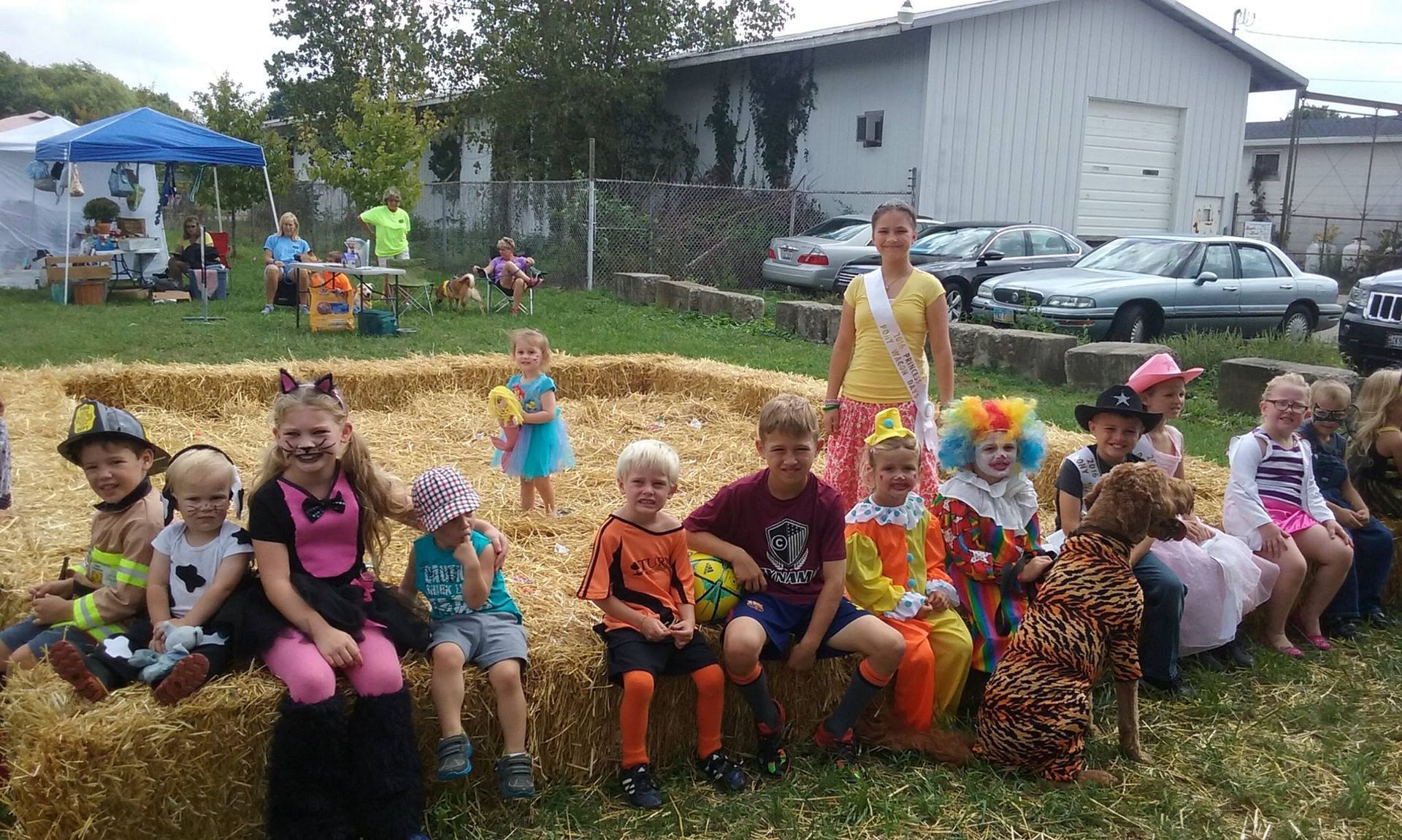 Children in costumes sit on hay bales at an outdoor event. A woman in sash stands nearby.