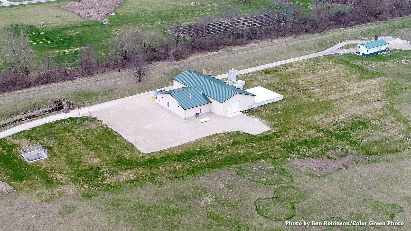 Aerial view of a light-colored building with a green roof next to a road, surrounded by grass and a water channel.