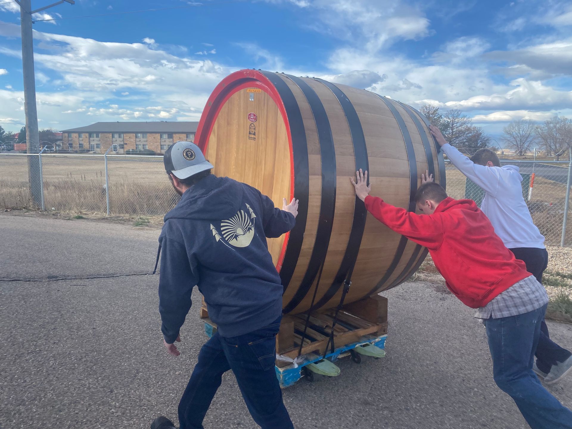 Two men are pushing a large wooden barrel down a road.