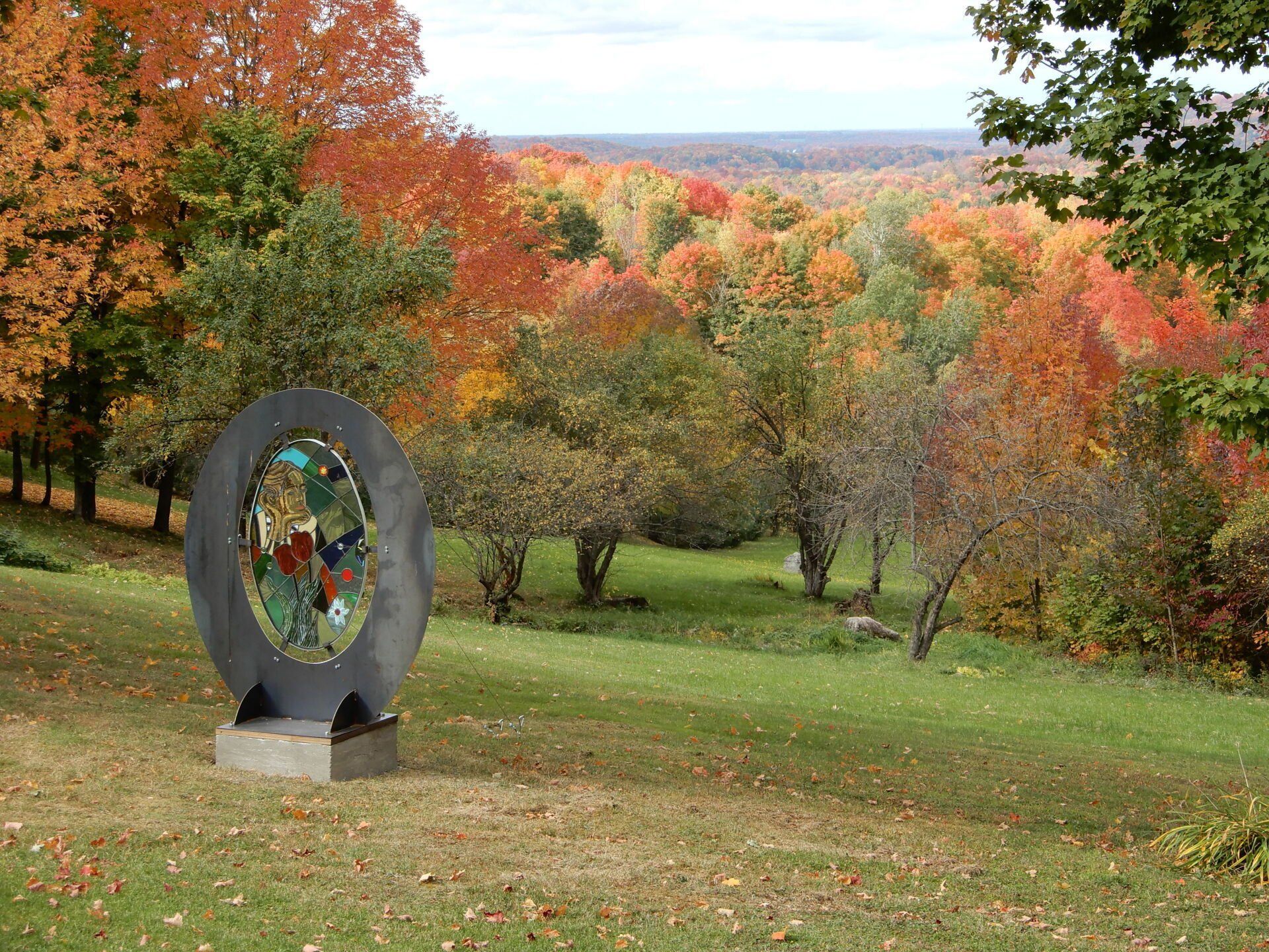 Sculpture on a grassy hill overlooking a forest with vibrant autumn foliage.