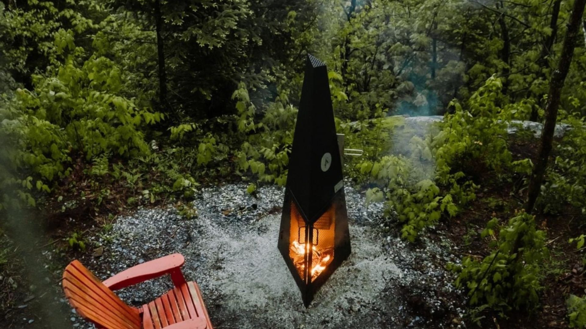 A tall, black fire pit burning in a lush green forest, with an orange chair in the foreground.