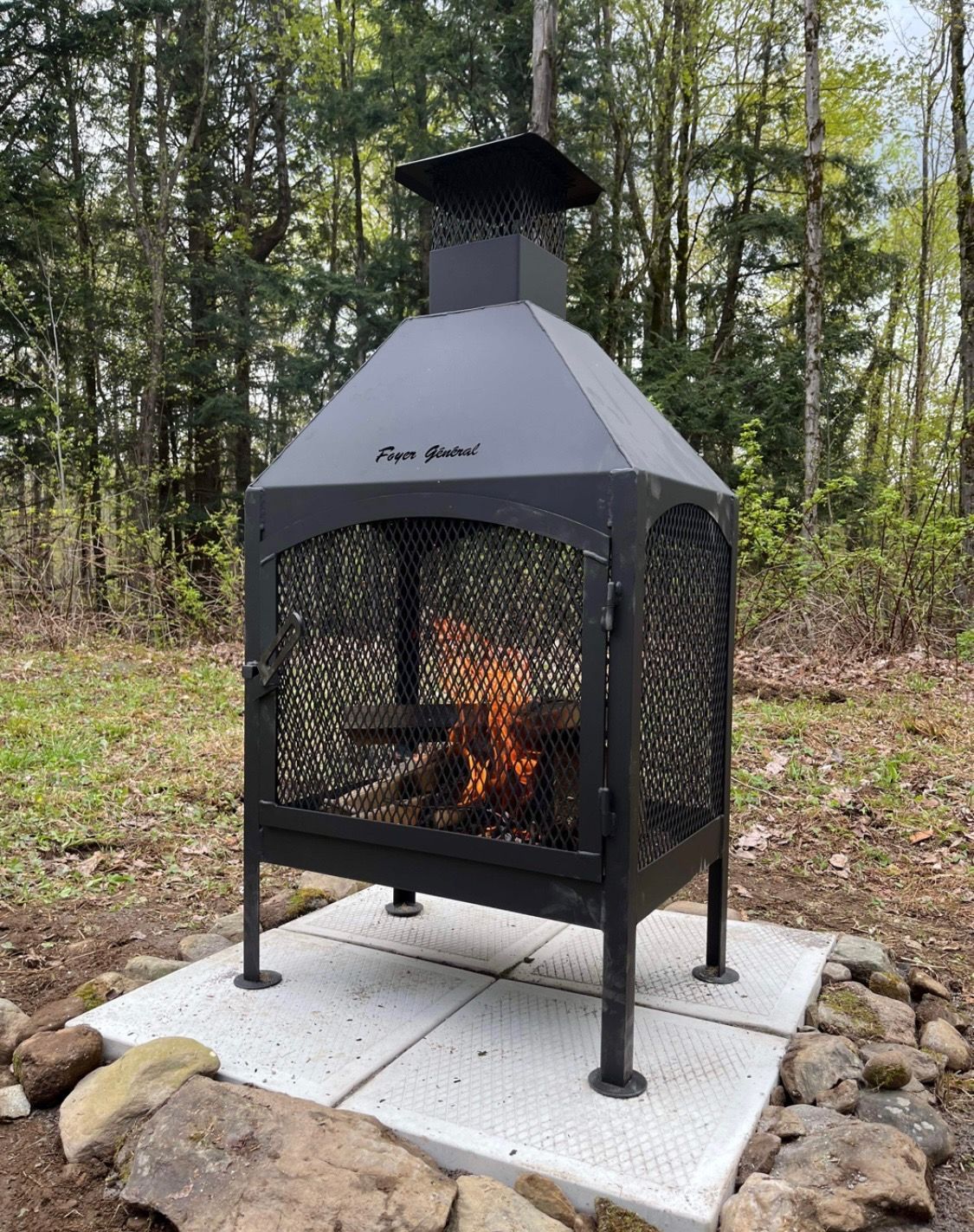 Black outdoor fireplace with flames burning, on a concrete slab surrounded by rocks, in a wooded area.
