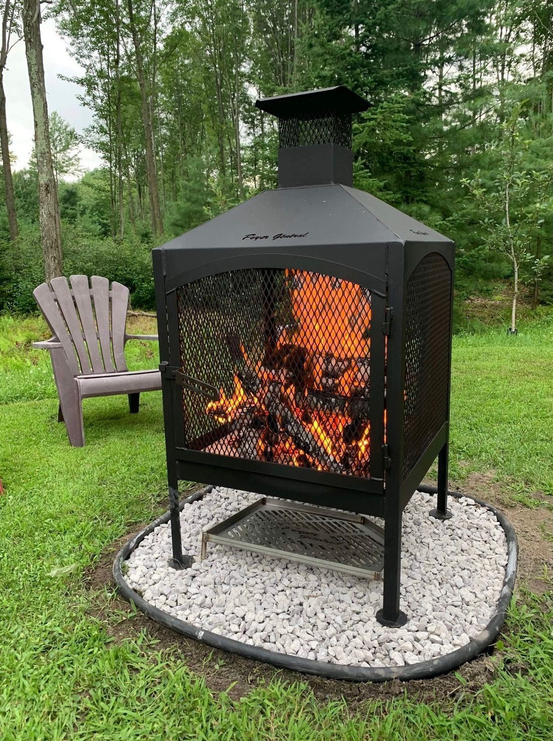 A lit black fire pit with mesh sides sits on a bed of rocks, next to a purple Adirondack chair on a grassy lawn.