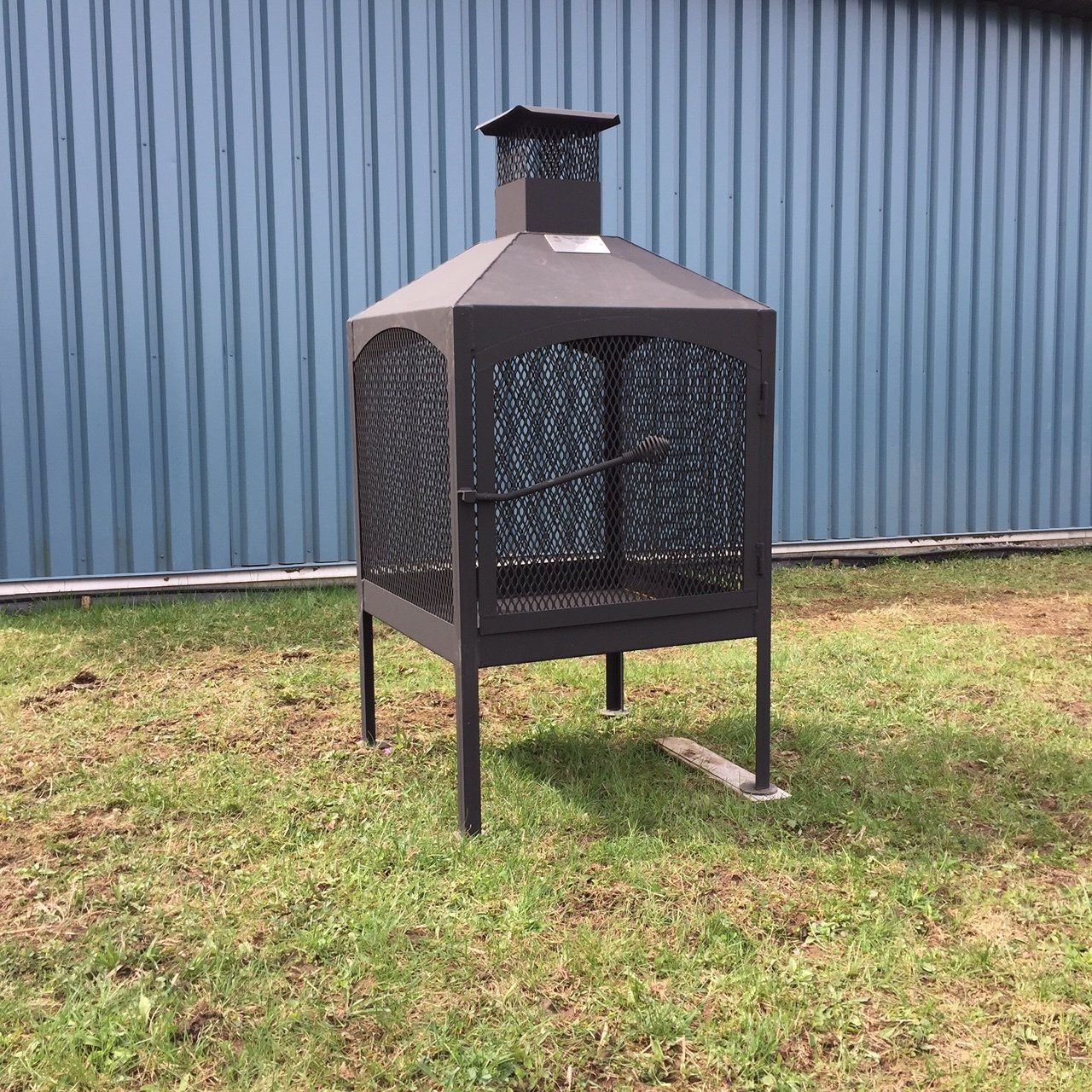 Black metal outdoor fireplace on grass against a blue building.