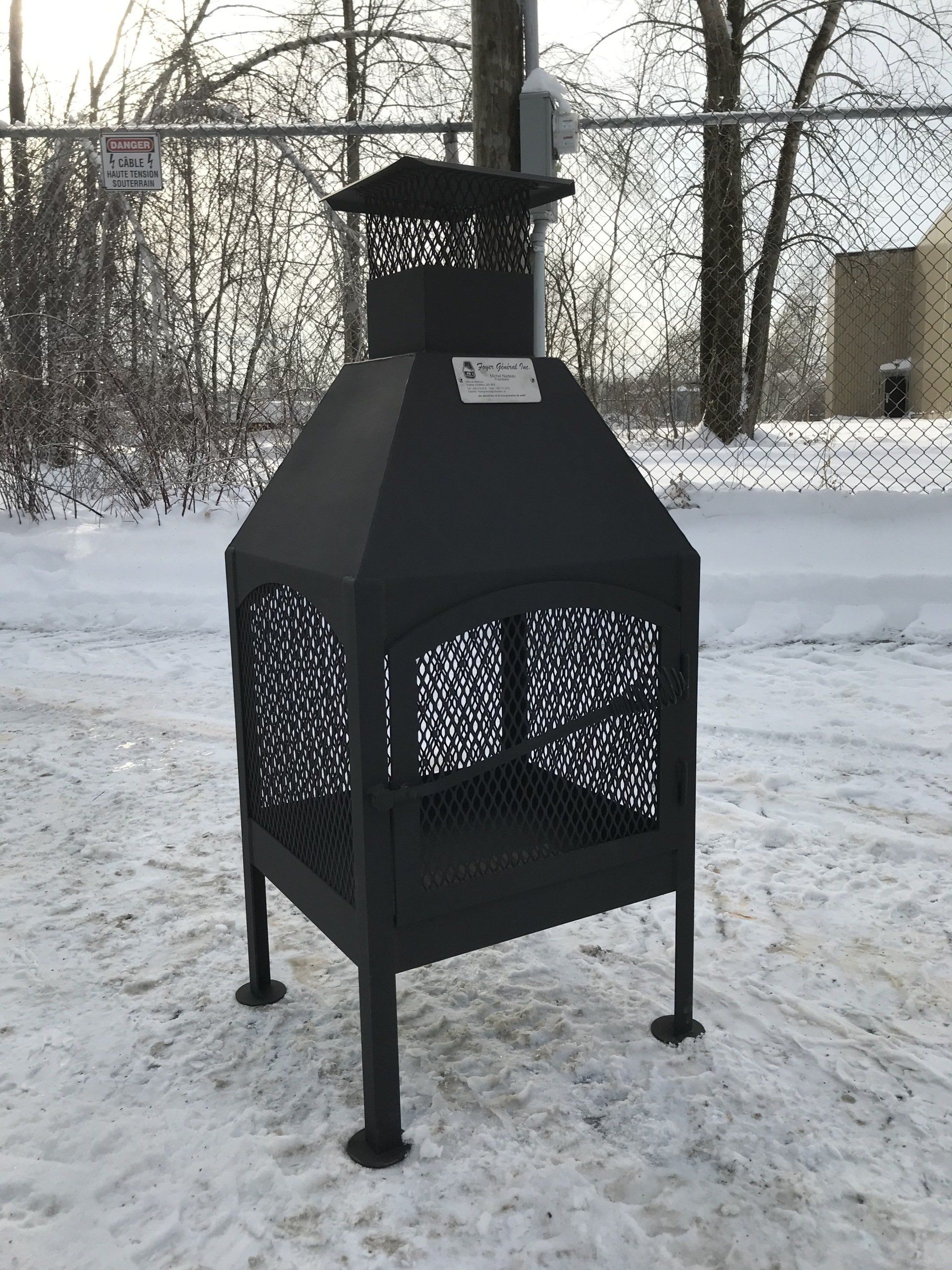 Black metal fire pit with a chimney, on a snowy ground, by a fence.