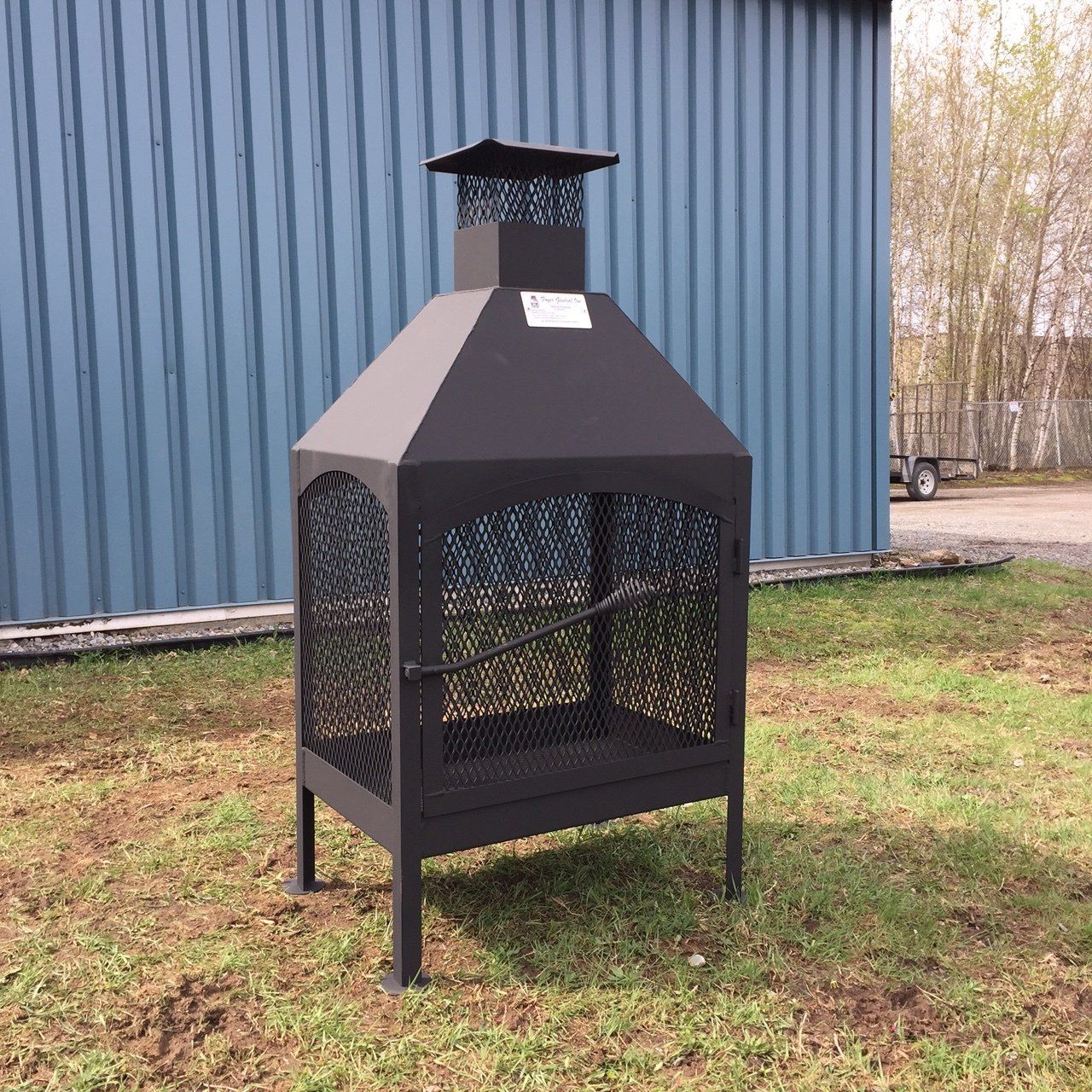Cheminée d'extérieur en métal noir avec côtés en filet, devant un bâtiment bleu sur une pelouse herbeuse.