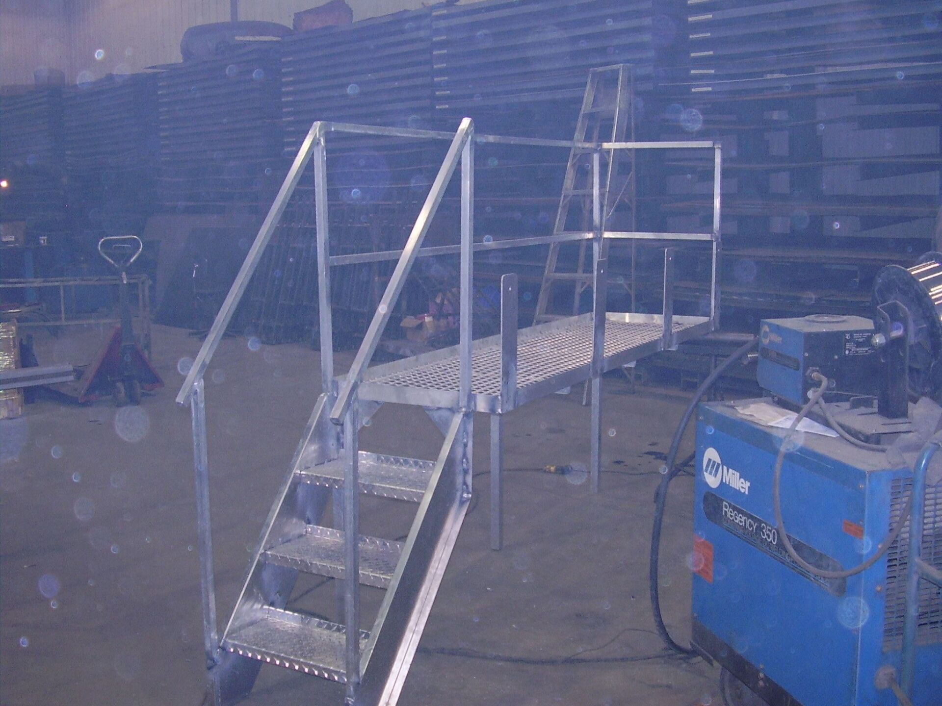 Metal platform with stairs and handrails next to a blue Miller welder in a warehouse setting.