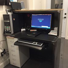 Un ordinateur est installé sur un bureau sombre. On y voit un ordinateur de bureau, un écran, un clavier et une imprimante. Des tiroirs blancs se trouvent en dessous.