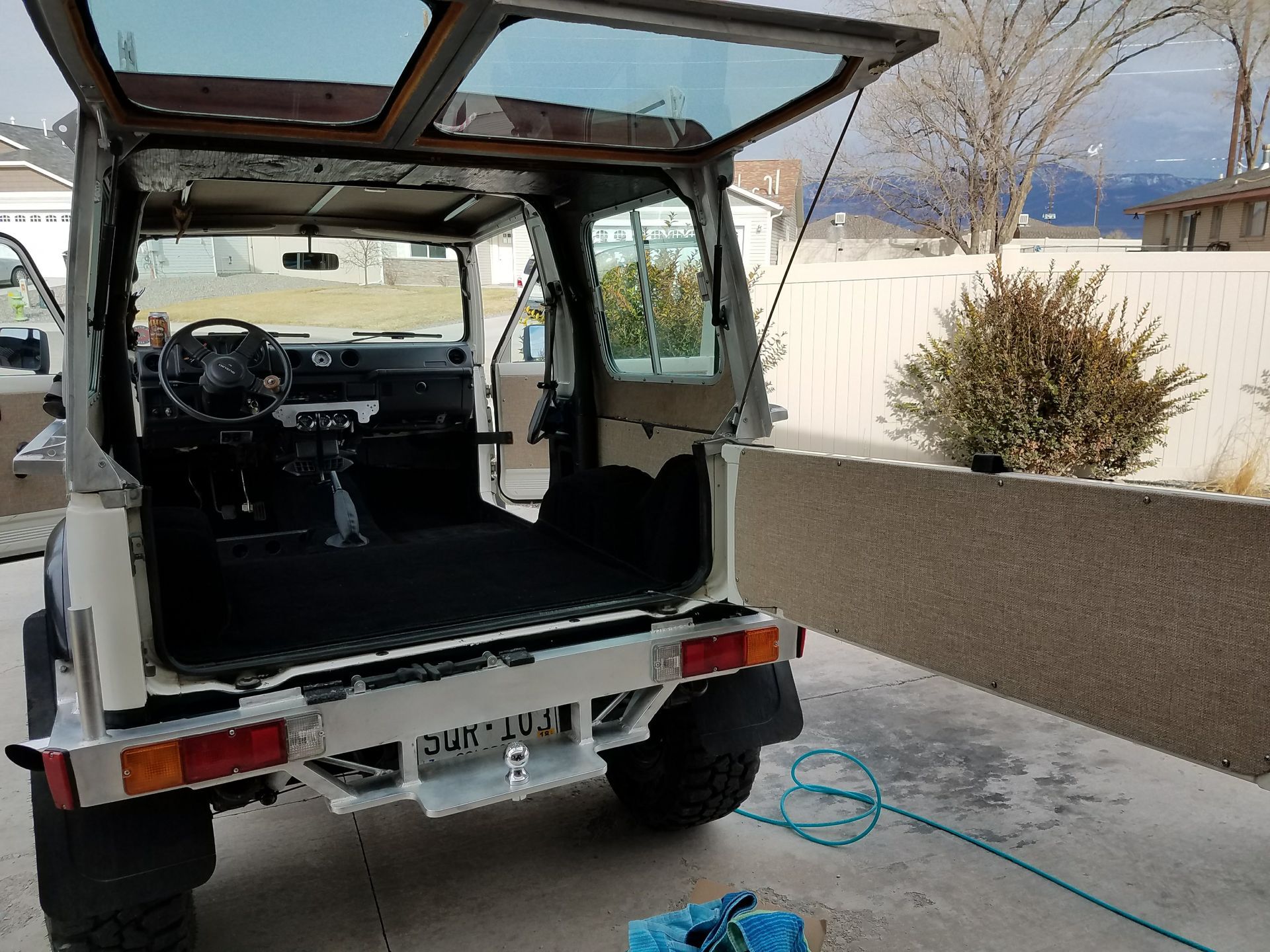 White Jeep with open rear hatch parked outdoors on a driveway, with mountains in the background.