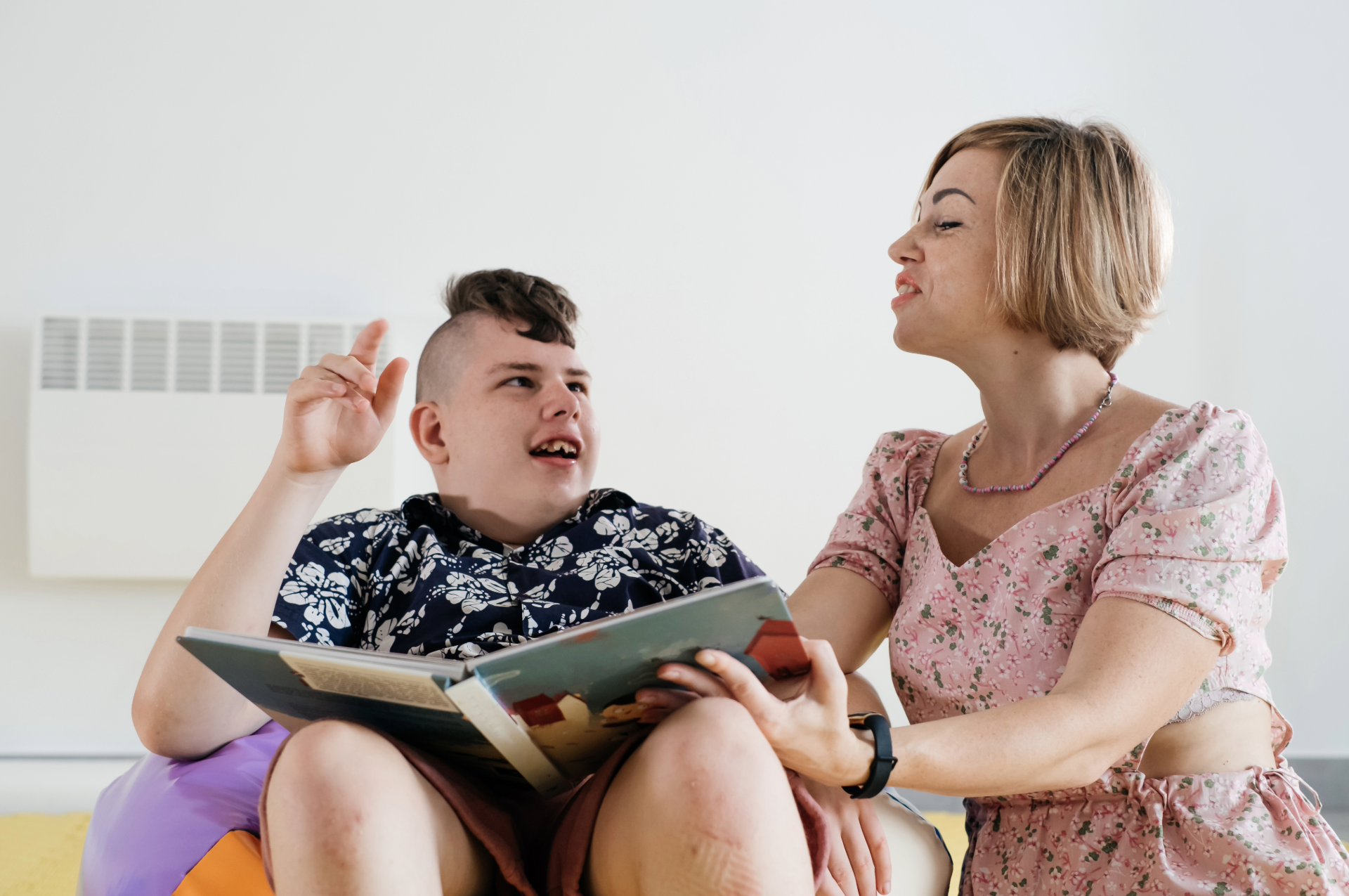 Woman helping a young man read a book in a brightly lit room.