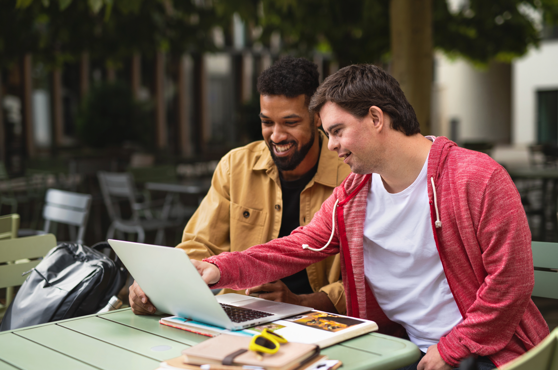 Two men look at a laptop outdoors. One points, smiling; the other smiles, wearing a red hooded sweatshirt.