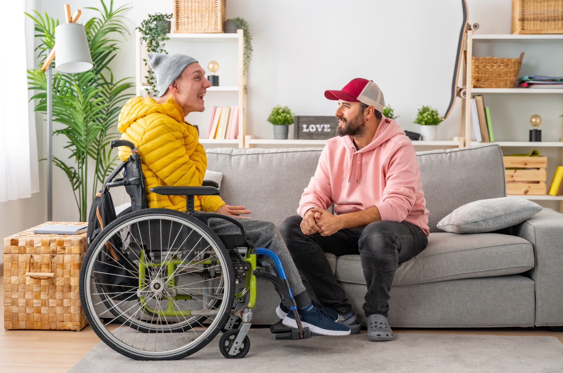 Two men in a living room, one in a wheelchair, talking. Man in wheelchair wears yellow jacket, gray hat. Man on couch wears pink hoodie, cap.