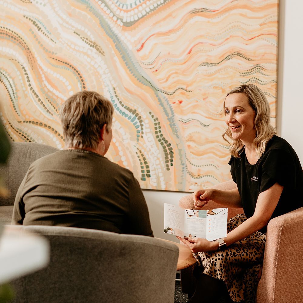Two women are sitting in chairs talking to each other in front of a painting.