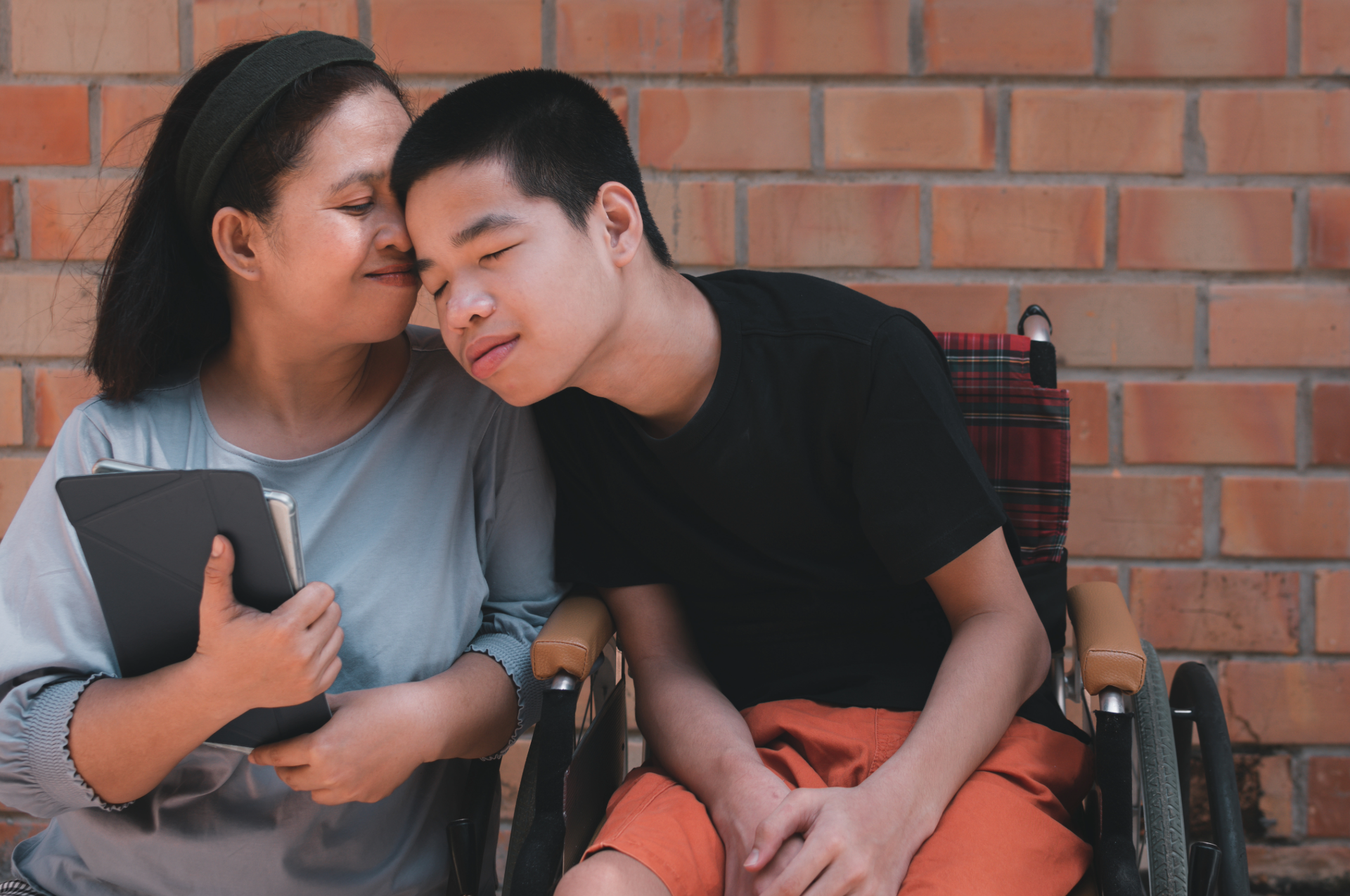 Woman gently embraces a young man in a wheelchair, both smiling softly, leaning against a brick wall.