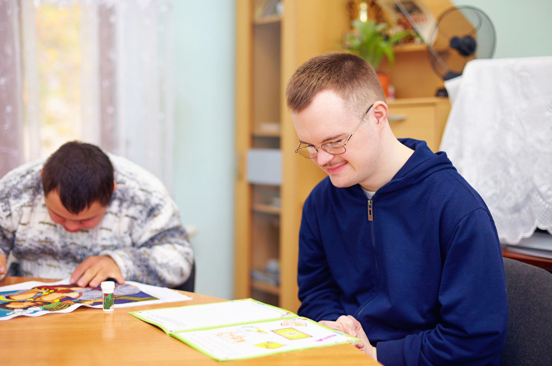 Two men with Down syndrome at a table, one reading and smiling, the other drawing. Indoors, warm lighting.