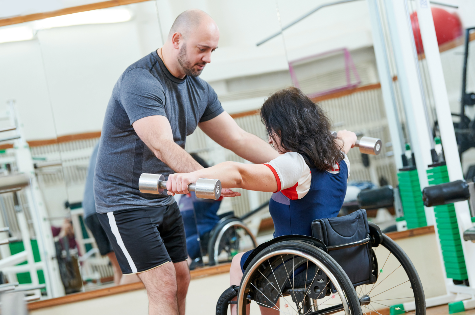 A man assisting a person in a wheelchair lifting dumbbells in a gym.