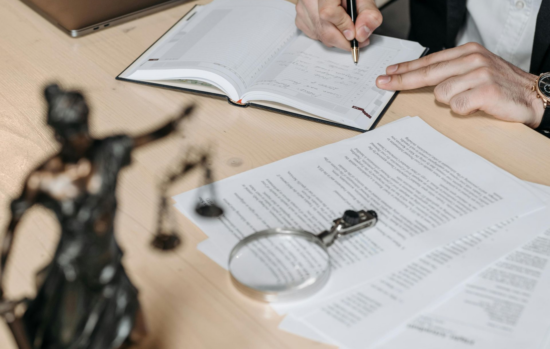 Person writing in notebook, legal documents, magnifying glass, and Lady Justice statue on desk.