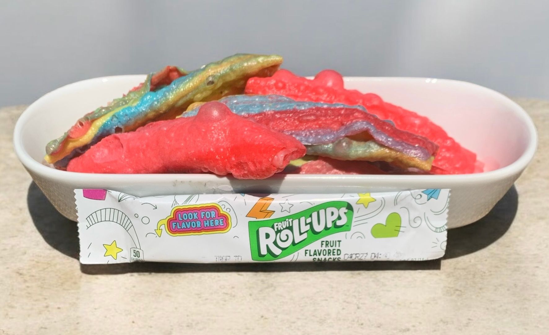 A bowl of gummy worms with a package of roll ups next to it.