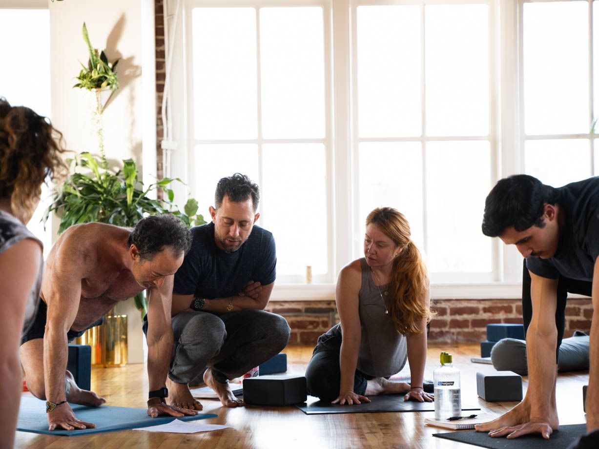People practicing yoga on mats in a bright studio, leaning forward in a seated pose.