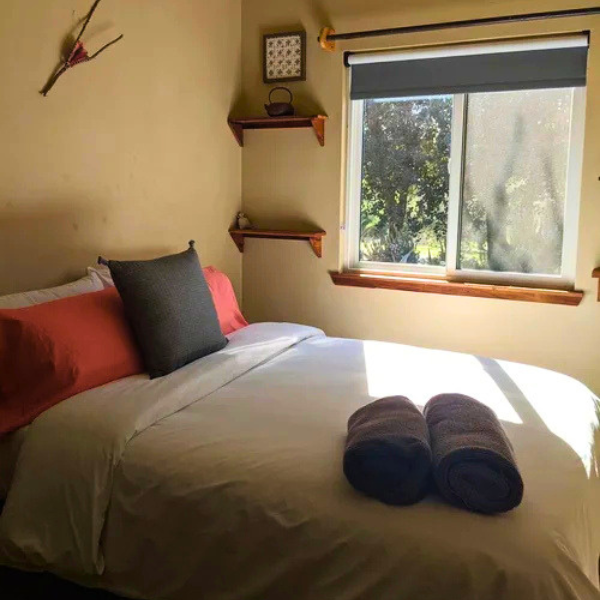 Cozy bedroom with a made bed, gray bedding, red pillows, rolled towels, and a sunlit window