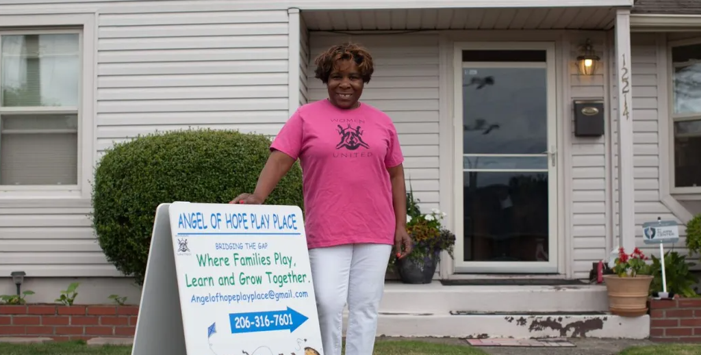 A woman in a pink shirt is standing in front of a house holding a sign.