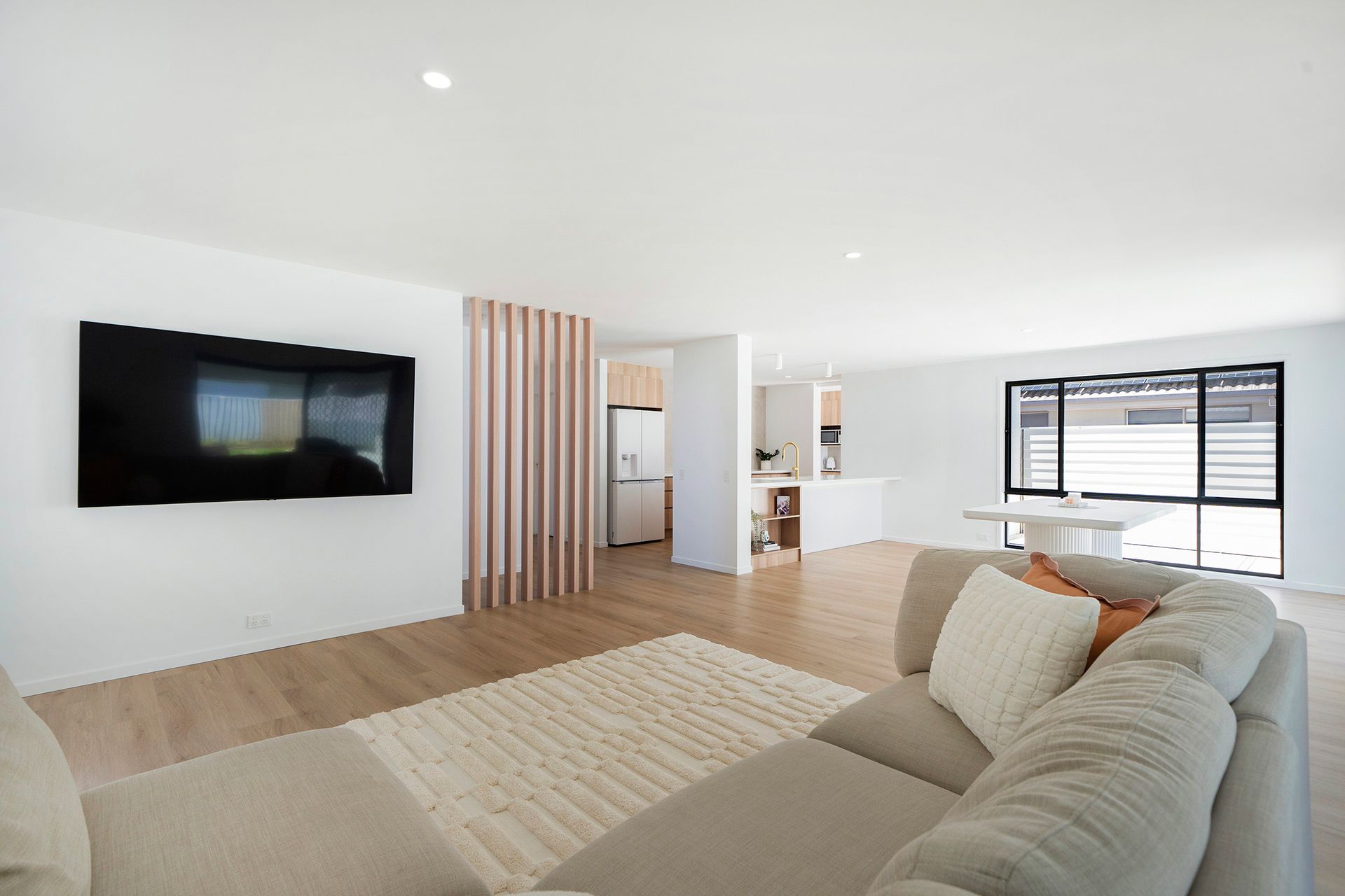 Bright modern living room with wall-mounted TV, beige sectional sofa, light wood floors, and kitchen area. — Reynolds Built In Caloundra, QLD