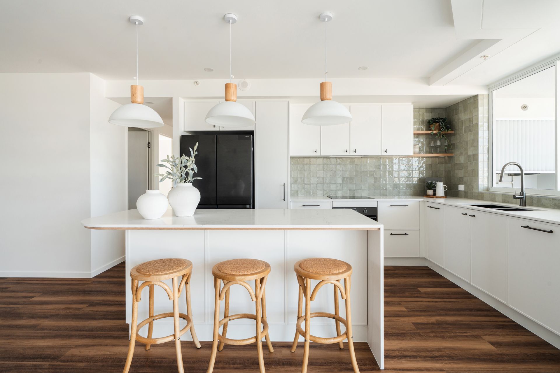 A modern kitchen featuring white cabinets, a large island with three rattan stools, and pendant lights over dark flooring.