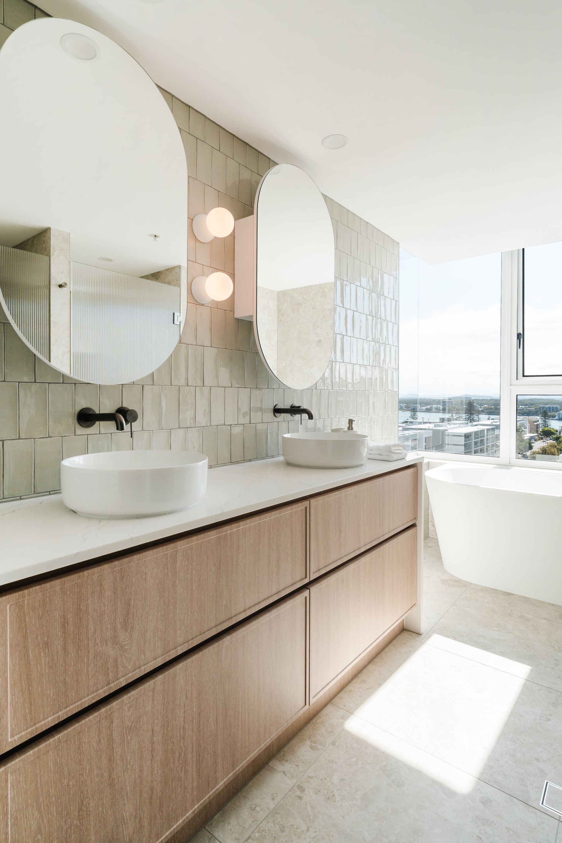 A modern bathroom vanity with two round vessel sinks, wooden cabinets, and white countertops against tiled walls.