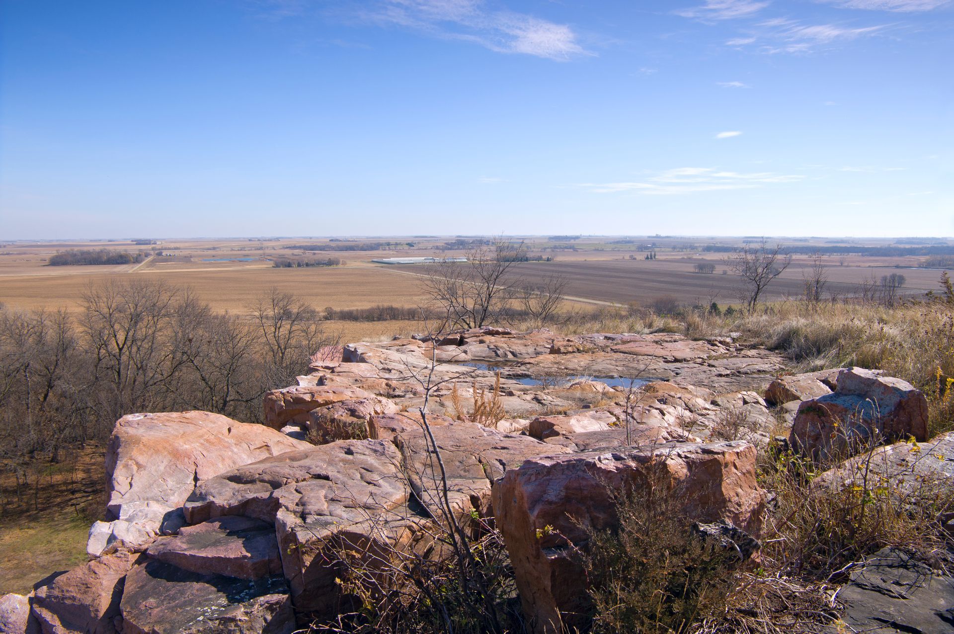 Scenic view from a rocky hilltop overlooking a flat, expansive landscape under a clear, blue sky.