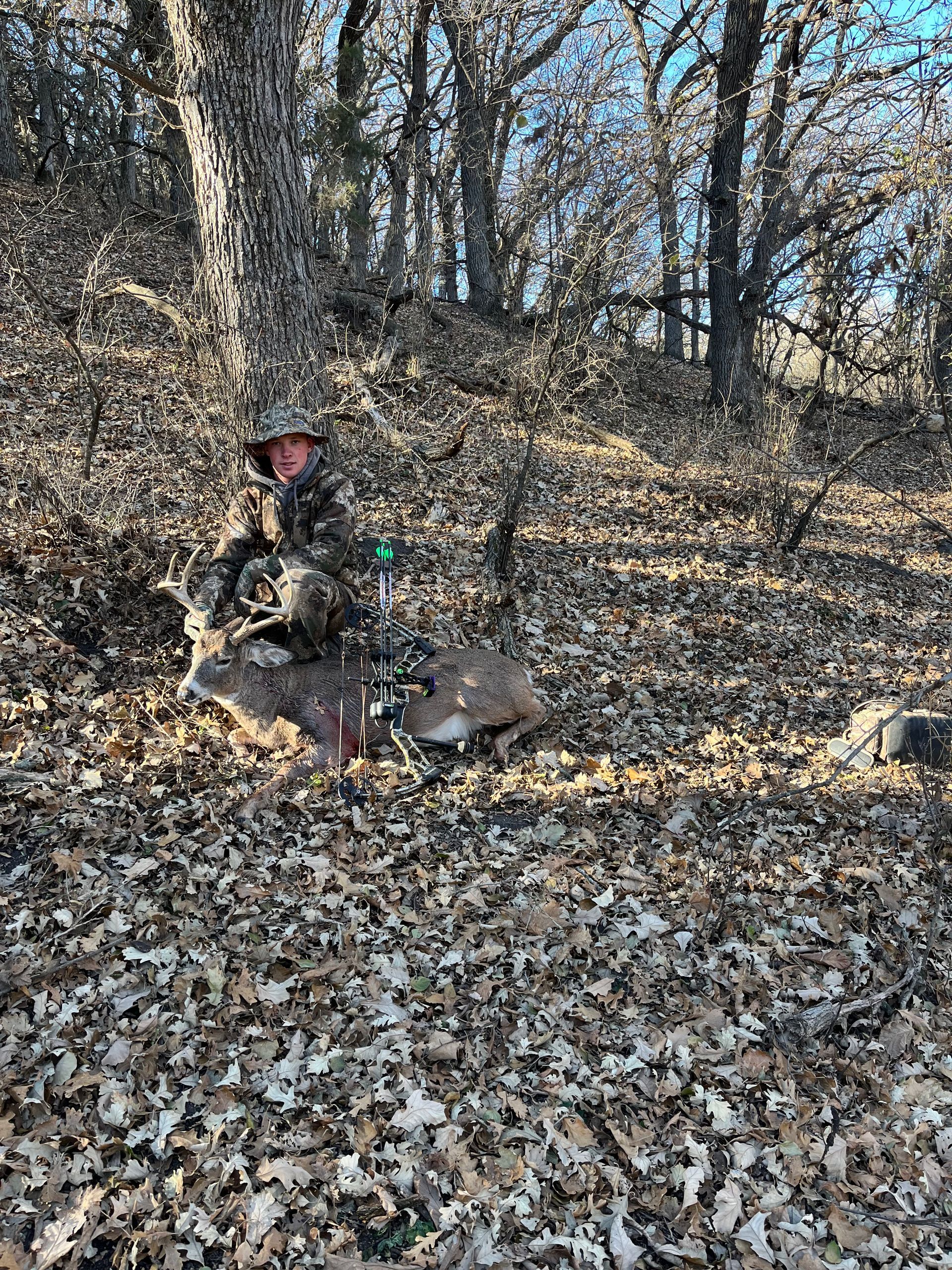 Hunter in camouflage kneels by a dead deer in a forest covered in leaves.