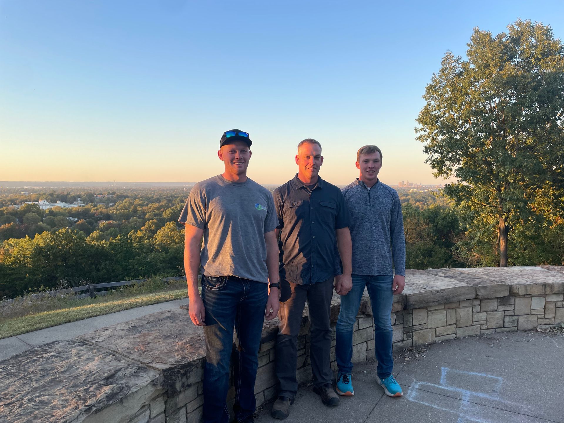 Three men stand on a stone wall overlooking a city at dusk.