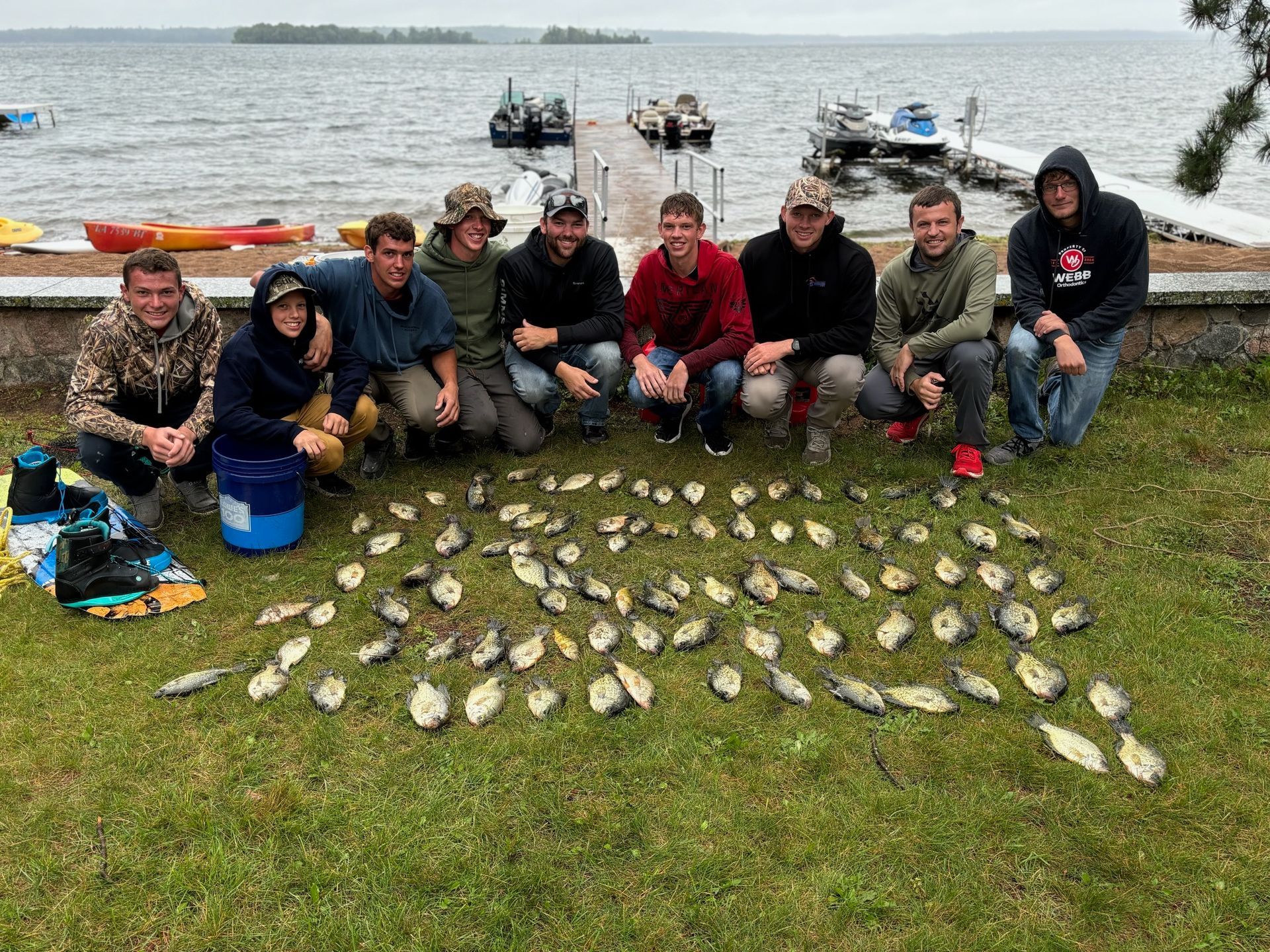 Group of men kneel with a large pile of fish on grass near a lake with boats and docks.