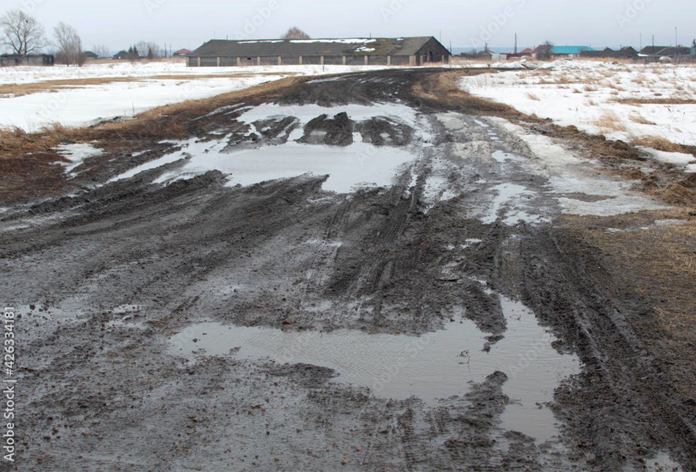 Muddy dirt road with puddles, snow patches, and a wooden barn in the distance.