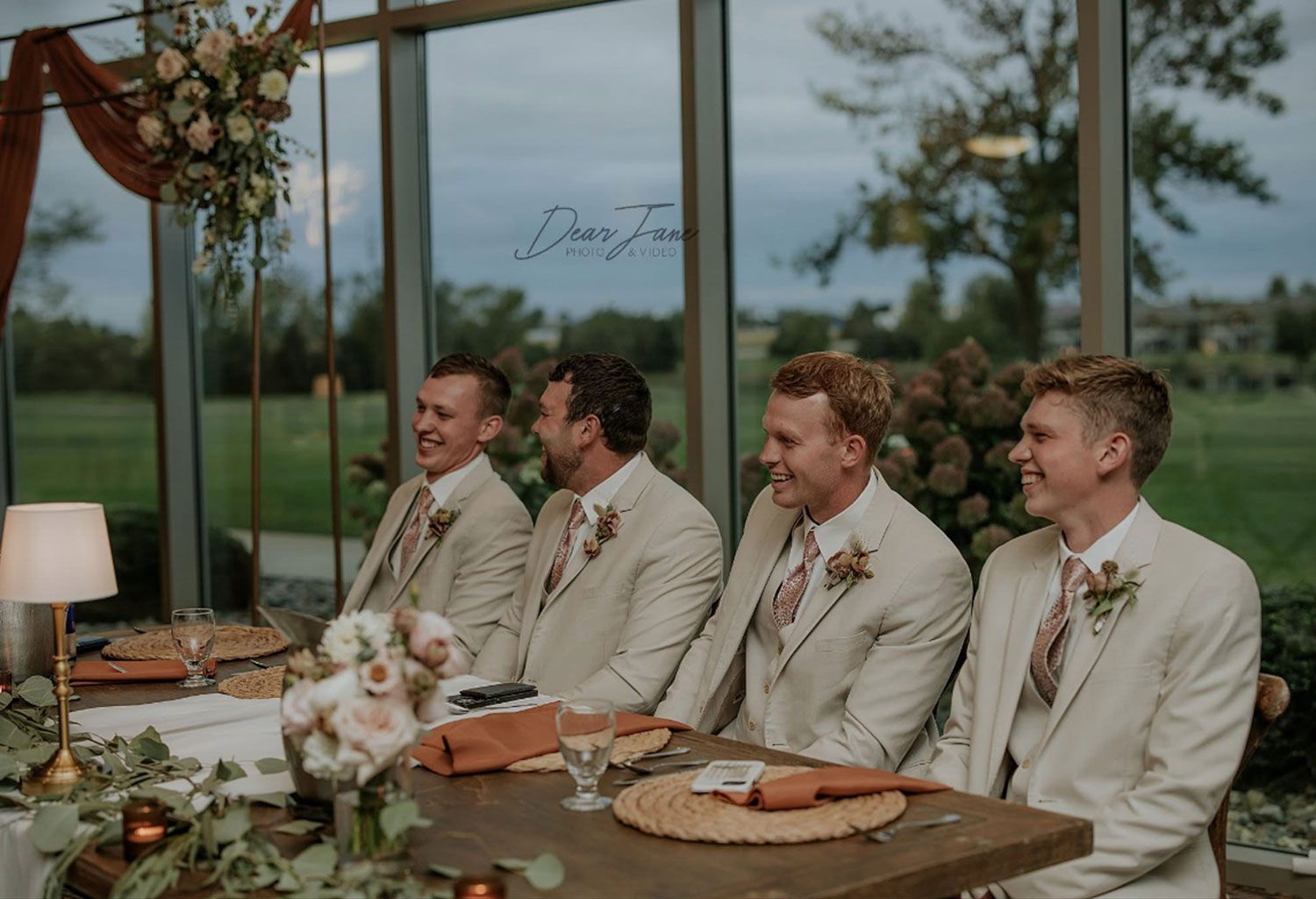Four men in tan suits smile at a wedding reception. They are seated at a table with decorations near a window.