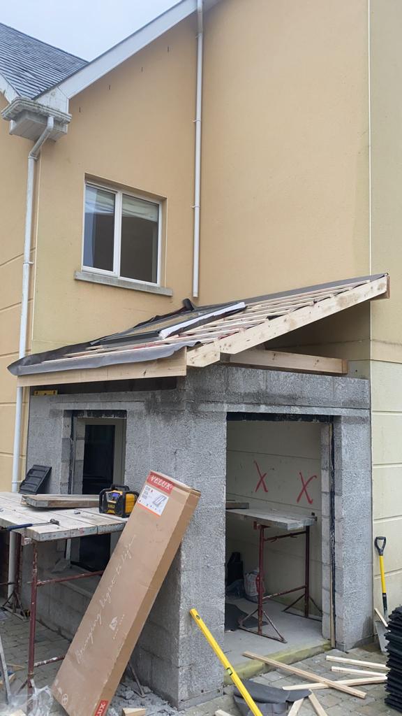 Construction of a roof extension on a cinder block structure adjacent to a beige house; tools and materials are visible.