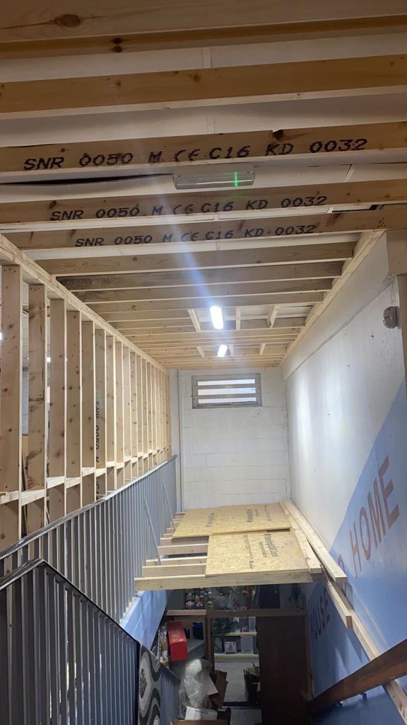 Stairwell construction with exposed wooden beams, studs, and plywood. Fluorescent lights and a grated window are visible.