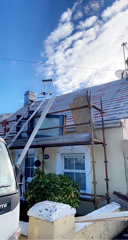 Building with scaffolding, undergoing roof work on a sunny day. Blue sky, white clouds, and visible roof tiles.