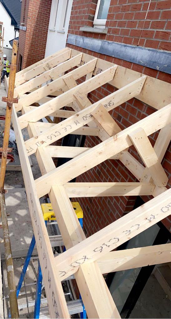 Wooden roof structure being built against a red brick building; scaffolding and a worker in the background.