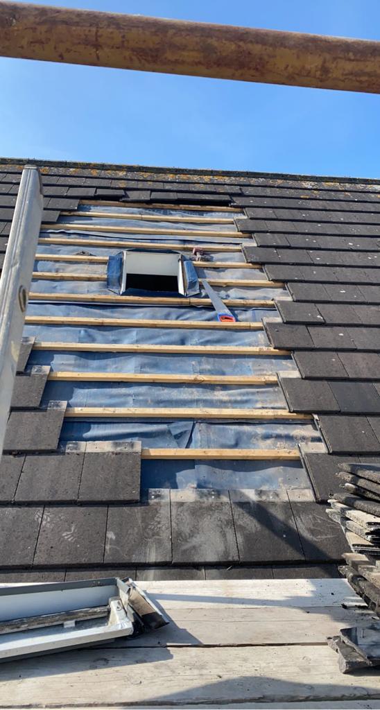 Roof partially torn off, revealing underlayment and rafters. A small square vent is visible. Blue sky in the background.