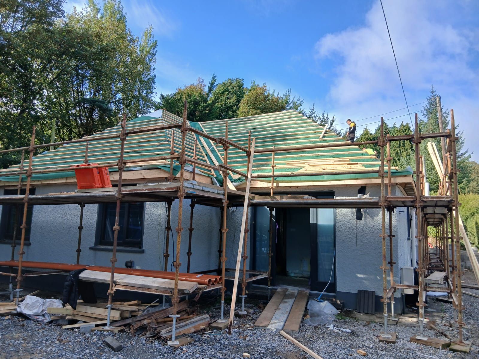 House under construction; scaffolding, green roof tiles, person on roof, trees in background, sunny day.