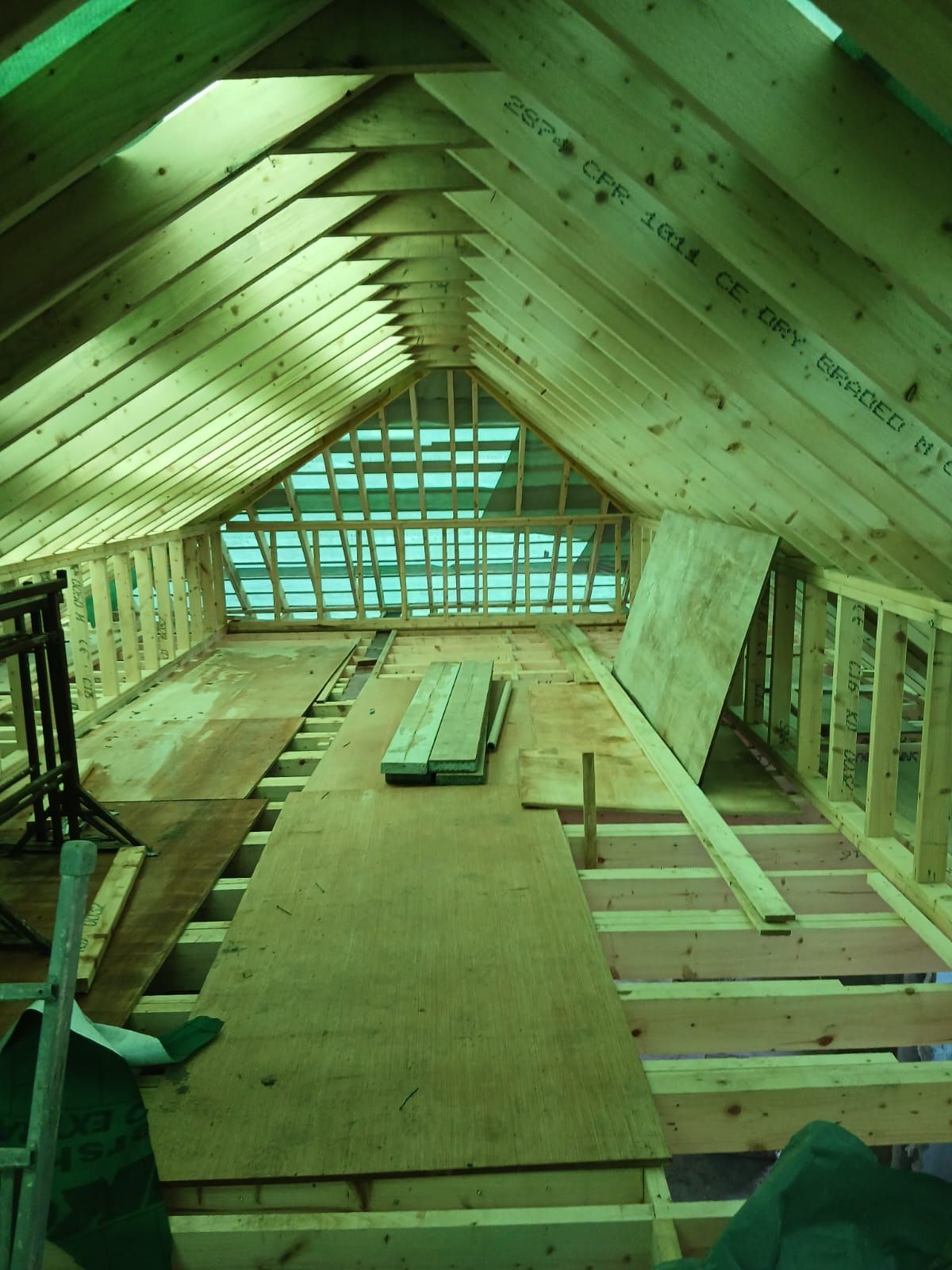 Interior view of a building's attic under construction; exposed wooden beams, plywood floor, and a triangular window.