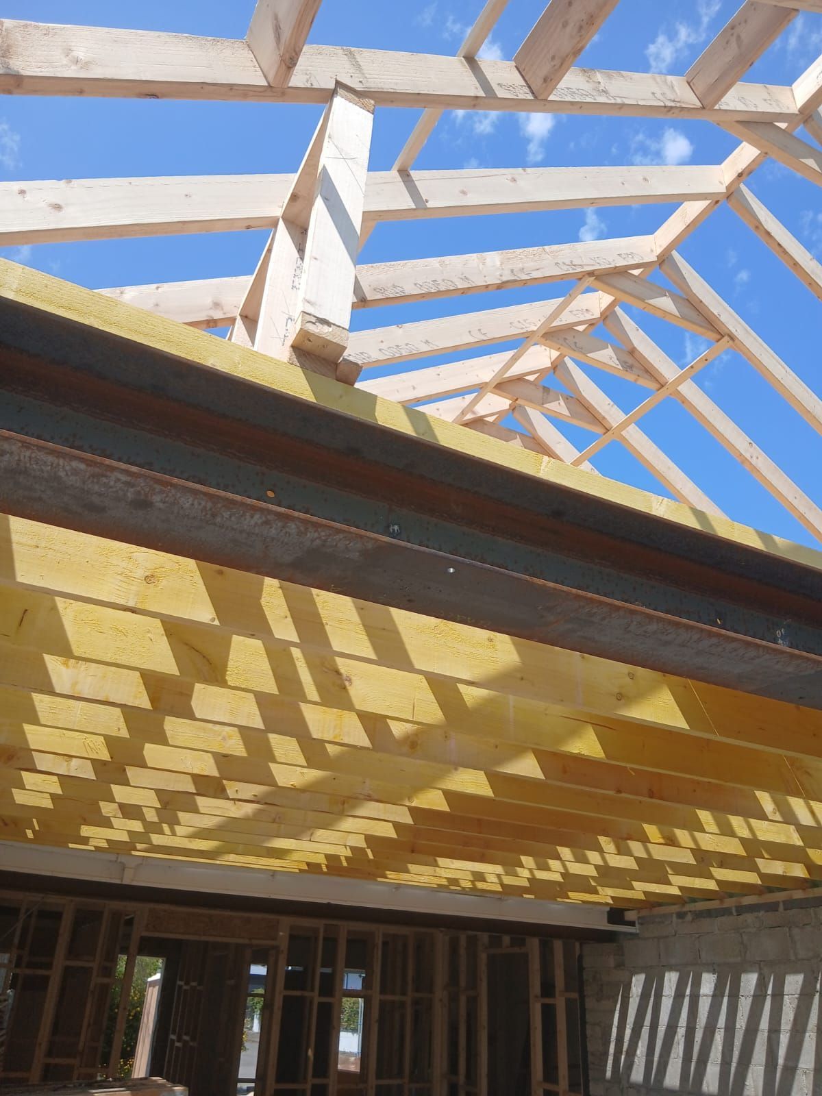 Construction of a roof structure. Wooden beams and rafters against a blue sky, with interior framing visible below.