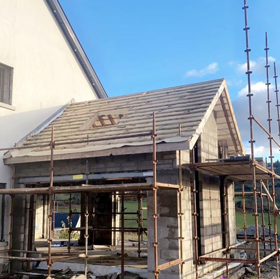 Construction of a small extension to a house. Scaffolding surrounds the unfinished brick structure under a slate roof.