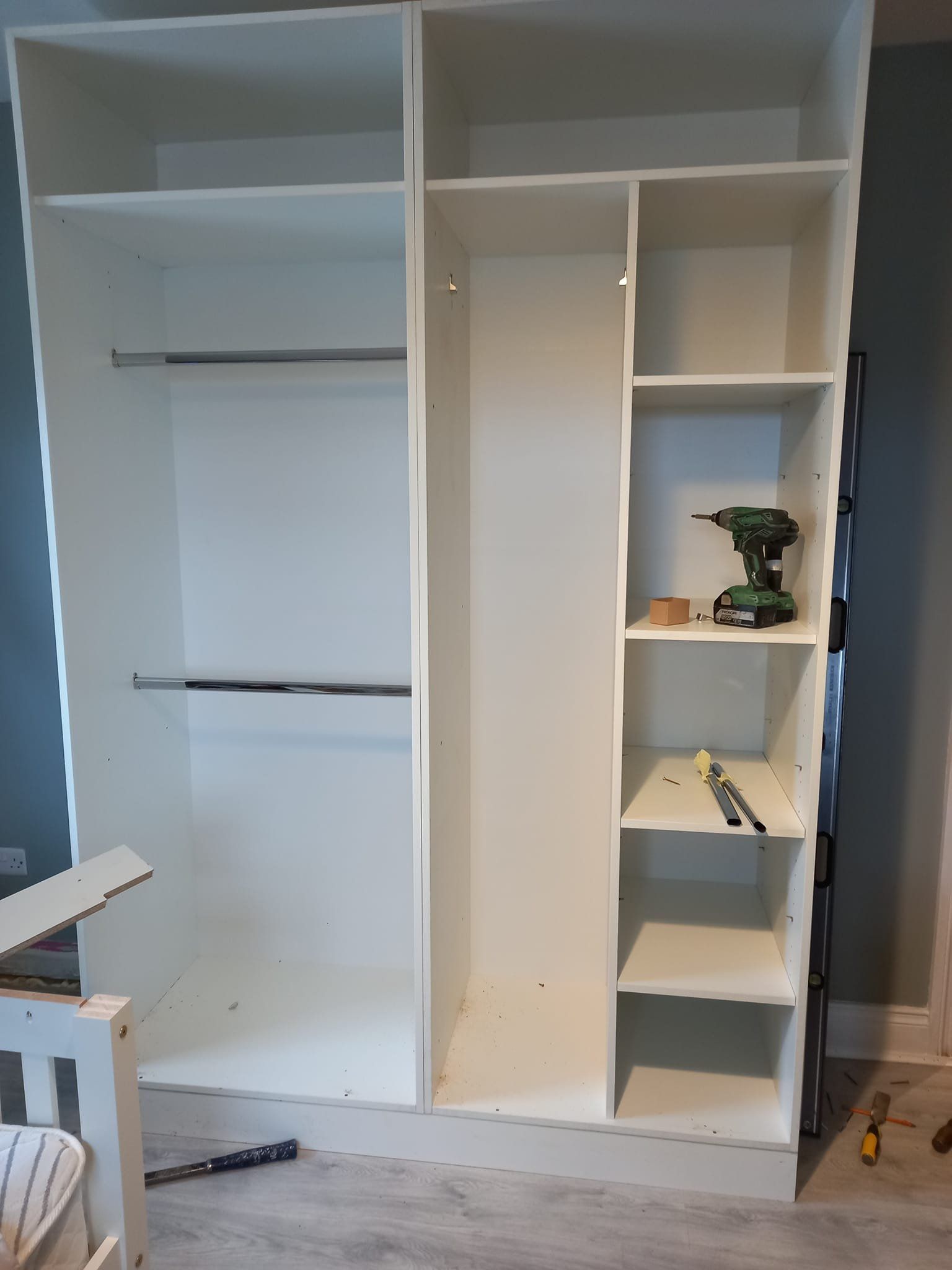 White wardrobe unit with shelves, hanging rods, and a power drill inside.
