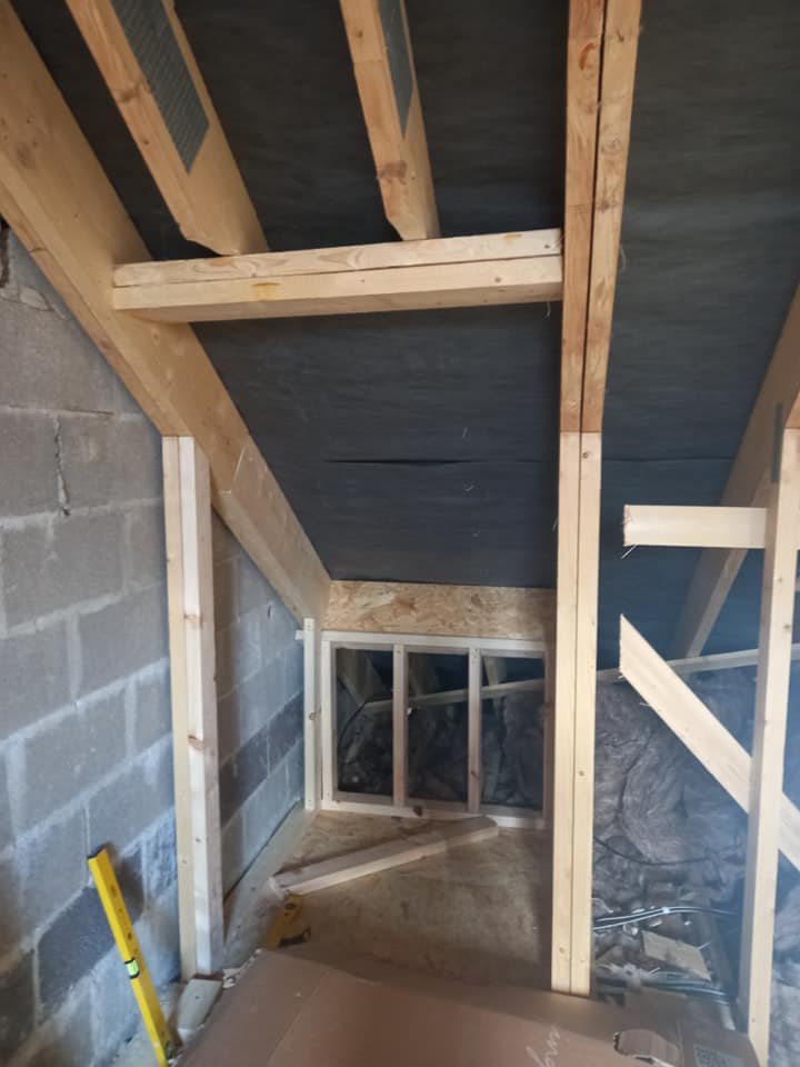 Interior view of an unfinished attic space with wooden framing and a window.