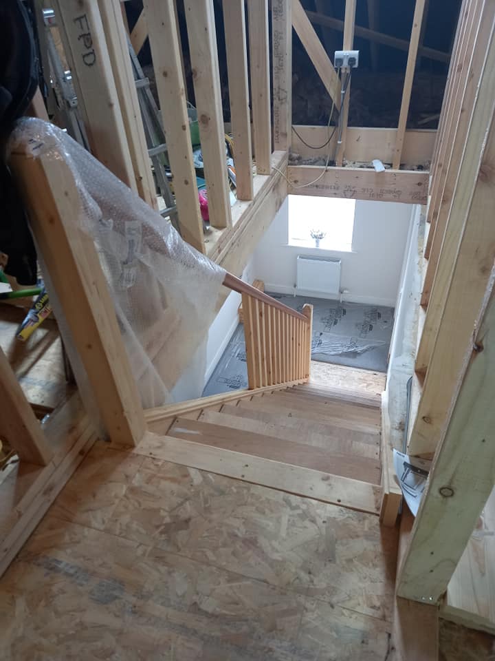 Construction site with wooden stairs leading down. Framing, plywood, and a partial railing are visible.