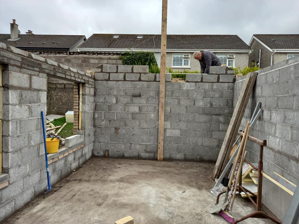 Construction of a concrete block building. A person works on the wall, a wooden pole is in the center.